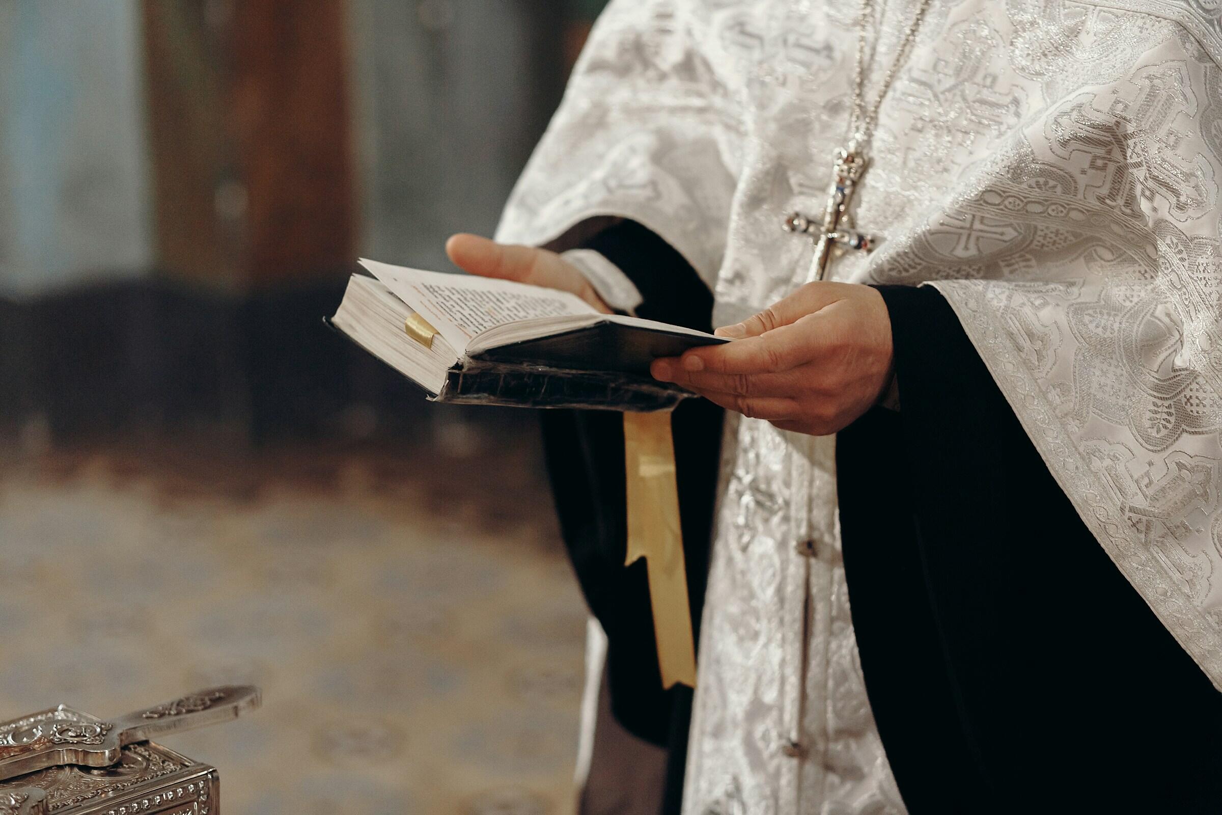 Un sacerdote sostiene un libro abierto con un texto religioso, vestido con una túnica blanca ornamentada, sobre un fondo ligeramente difuminado.