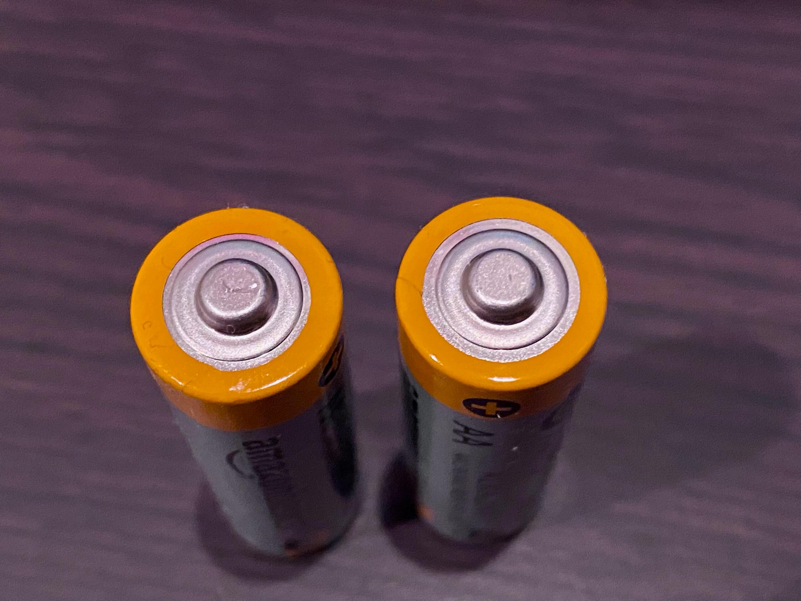 Dos pilas AA con la parte superior naranja colocadas en posición vertical sobre una superficie de madera oscura.